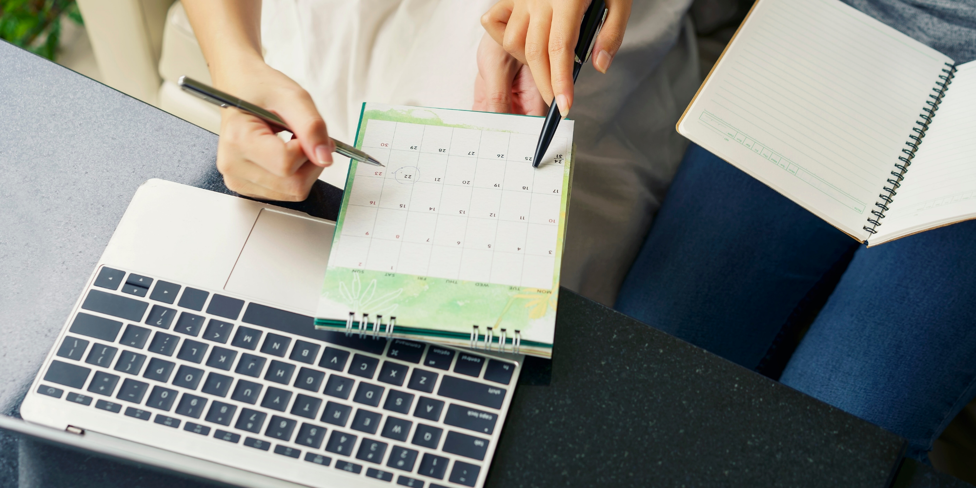 Two people mark dates on a paper calendar beside a laptop, using a digital planner app for group scheduling